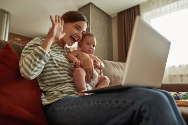 Mutlu aile çocuğu olan genç anne dizüstü bilgisayara bakıp el sallıyor.