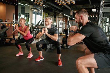 Aktif yaşlı kadın ve erkek spor salonunda çömelme hareketleri yapıyorlar.