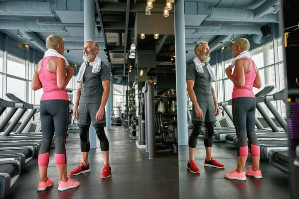 Sporty aged woman and man in gym, mirror reflection of senior couple