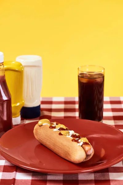 Cachorro quente acabado de fazer com mostarda e ketchup em uma placa com vidro de coque em pano de mesa de gingham vermelho sobre fundo amarelo — Fotografia de Stock