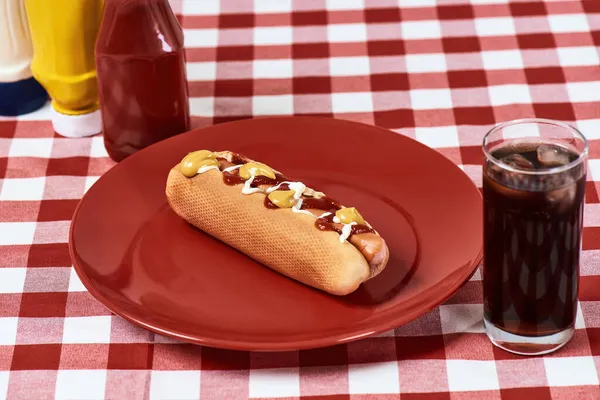 Closeup de cachorro-quente saboroso com mostarda e ketchup em uma placa com vidro de coque sobre guardanapo xadrez xadrez vermelho — Fotografia de Stock