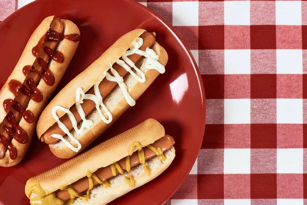 Flatlay de cachorros-quentes saborosos com mostarda e ketchup em uma placa sobre guardanapo xadrez xadrez vermelho — Fotografia de Stock