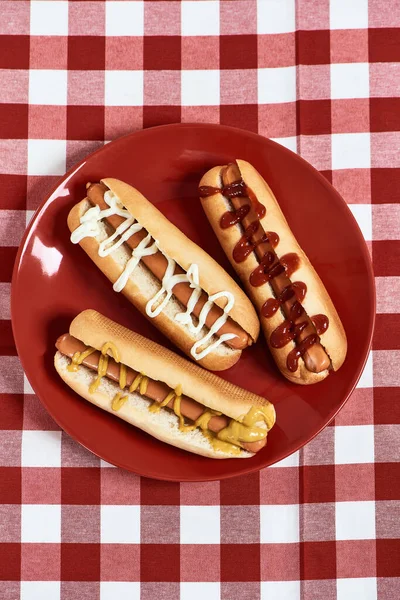 Vista superior de cachorros-quentes recém-feitos com mostarda e ketchup em um prato sobre toalha de mesa de gingham vermelho — Fotografia de Stock