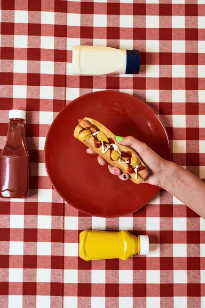 Close up of female hand holding freshly made hot dog with mustard and ketchup over red gingham table cloth — Stock Photo, Image