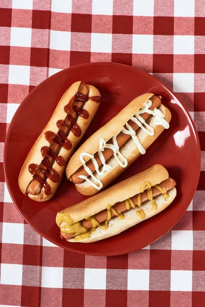 Vista superior de cachorros-quentes saborosos com mostarda e ketchup em uma placa sobre guardanapo xadrez xadrez vermelho — Fotografia de Stock