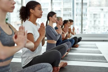 Çeşitli kadınlar kulüpte sıra sıra oturup yoga çalışırlar.