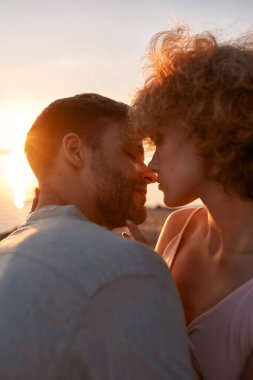 Aşık genç çift öpüşmeden önce gözlerini kapatıyor. Gün batımında kumsalda vakit geçiriyorlar.