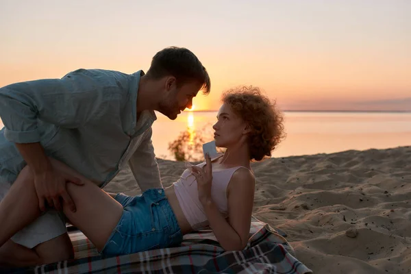 Beau jeune couple allongé sur la plage au coucher du soleil, prêt à faire l'amour. Femme tient un préservatif — Photo