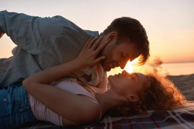 Romantik genç çift sahilde öpüşüyor, arkalarında güneş varken, gün batımında kumsalda birlikte uzanıyorlar.