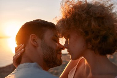 Gün batımında sahilde vakit geçirirken öpüşmeden önce gözlerini kapatıp birbirlerine dokunan genç çiftin portresini kapat.