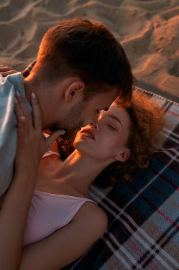 Gün batımında plajda birlikte uzanırken, birbirine aşık genç çift öpüşüyor ve sarılıyor.