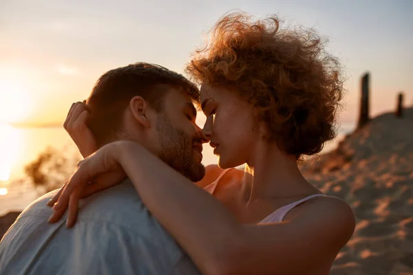 Retrato de jovem casal apaixonado fechando os olhos antes do beijo, sentado na praia ao pôr do sol — Fotografia de Stock