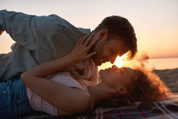 Casal jovem romântico beijando na costa com o sol atrás deles, deitados juntos na praia ao pôr do sol — Fotografia de Stock