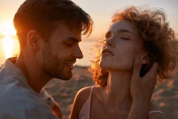 Retrato de jovem casal apaixonado parecendo apaixonado, tocando uns aos outros enquanto passa o tempo na praia ao pôr do sol — Fotografia de Stock