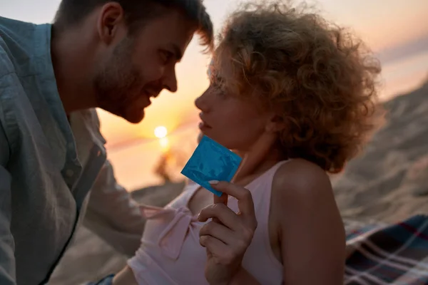 Portrait de jeune couple prêt pour faire l'amour sur la plage au coucher du soleil. Femme tient et montre un préservatif à la caméra — Photo