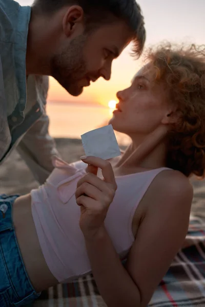 젊은 부부는 해 가 지면 해변에서 사랑을 나눌 것이다. 여자가 콘돔을 들고 카메라를 보여 주고 있습니다 — 스톡 사진