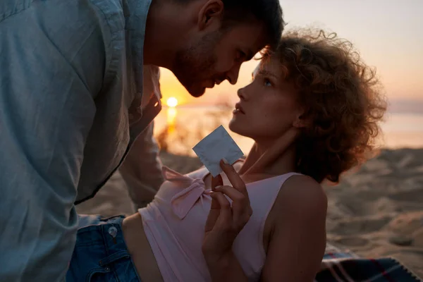 Belo jovem casal pronto para fazer amor na praia ao pôr do sol. Mulher está mostrando um preservativo para câmera — Fotografia de Stock