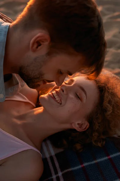 Feliz jovem casal apaixonado beijando e rindo enquanto deitado juntos na praia ao pôr do sol — Fotografia de Stock