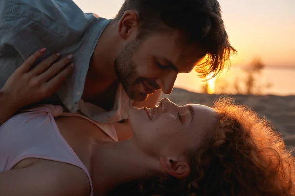 Romantic young people in love cuddling on the shore with the sun behind them, lying together on the beach at sunset — Stock Photo, Image