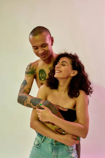 Mezza nuda giovane ragazzo tatuato e fidanzata con i capelli ricci scuri cercando felice mentre posa insieme isolato su sfondo luce al neon — Foto Stock