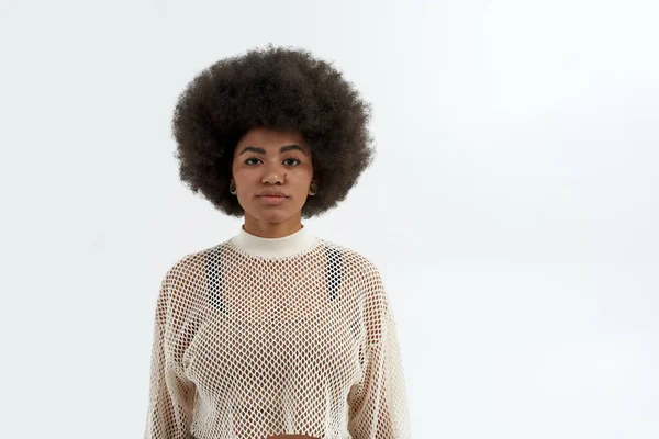 Genç, ciddi, siyah bir kadın kameraya bakıyor. Modern gençlik tarzı. Şık, çekici, kıvırcık saçlı, örgülü tişört giyen kız. Beyaz arka planda izole edilmiş. Stüdyo çekimi. Boşluğu kopyala