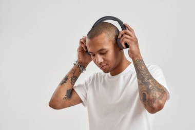 Karışık ırk dövmeli bir adamın portresi kulaklık takıyor, aşağı bakıyor, hip hop müzik dinlemeye hazır, beyaz arka planda soyutlanmış poz veriyor.