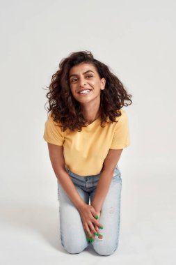 Siyah kıvırcık saçlı neşeli genç bir kadın. Neşeli görünüyor, gülümsüyor. Aydınlık arka planda yalnız poz veriyor.