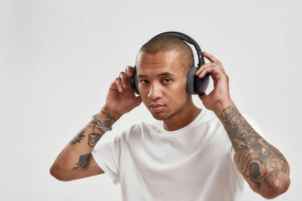 Kulaklık takan, kameraya bakan ve beyaz arka planda izole bir şekilde poz verirken hip hop dinleyen melez dövmeli bir adamın portresi. — Stok fotoğraf