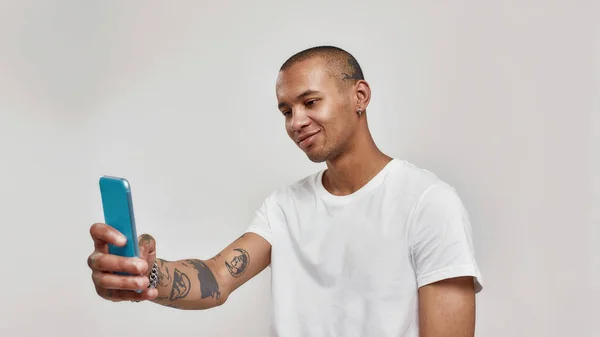Ritratto di giovane uomo tatuato di razza mista in t-shirt scattare selfie utilizzando smartphone mentre in piedi isolato su sfondo chiaro — Foto Stock