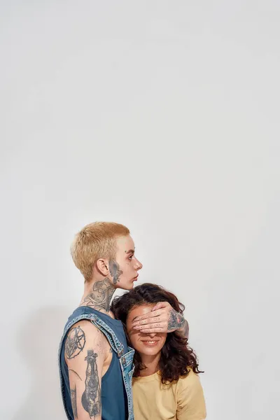 Due diverse giovani donne, coppia lesbica posa insieme isolato su sfondo grigio. Donna che copre i suoi occhi fidanzata con mano — Foto Stock