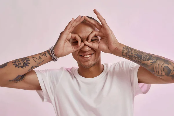 Retrato de jovem alegre tatuado misto raça cara no branco t-shirt sorrindo para câmera, fazendo óculos gesto com as mãos, posando isolado sobre rosa fundo — Fotografia de Stock