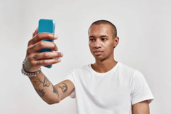 Ritratto di attraente tatuato misto razza giovane ragazzo in t shirt scattare selfie utilizzando smartphone mentre in piedi isolato su sfondo chiaro — Foto Stock