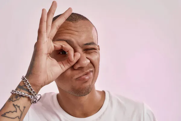 Retrato de jovem alegre tatuado misto raça cara no branco t-shirt fazendo ok gesto sobre olho, posando isolado sobre rosa fundo — Fotografia de Stock