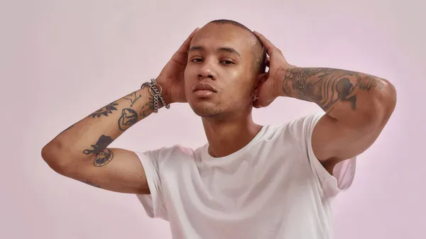 Retrato de confiante jovem mestiço tatuado cara em camiseta branca de mãos dadas em sua cabeça enquanto posando para câmera isolada sobre fundo rosa — Fotografia de Stock