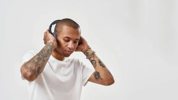Kulaklık takan, beyaz arka planda izole bir şekilde poz verirken hip hop dinleyen odaklanmış melez dövmeli genç bir adam. — Stok fotoğraf