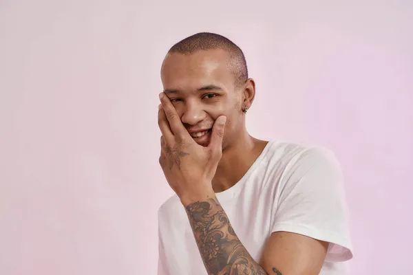 Retrato de feliz jovem mestiço homem tatuado em camiseta branca sorrindo para a câmera, tocando seu rosto, posando isolado sobre fundo rosa — Fotografia de Stock