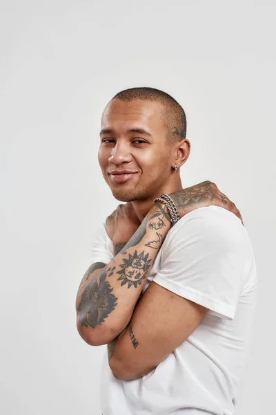 Portret van lachende jonge gemengde ras getatoeëerde man in wit t shirt hand in hand rond zijn nek, terwijl poseren voor camera geïsoleerd over witte achtergrond — Stockfoto