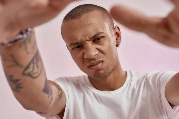 Közelkép elegáns fiatal vegyes rasszú tetovált srác fehér pólóban nézi a kamera, kinyújtja a karját, miközben pózol elszigetelt rózsaszín háttér — Stock Fotó