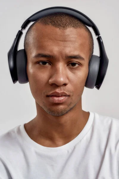 Close-up retrato de grave mestiço cara vestindo fones de ouvido olhando para câmera, ouvindo música ou podcast enquanto posando isolado sobre fundo branco — Fotografia de Stock