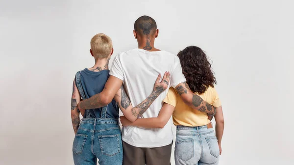 Vista posteriore di tre giovani amici diversi in abbigliamento casual che si abbracciano, in piedi insieme isolati su sfondo chiaro — Foto Stock
