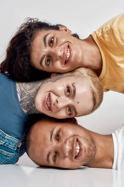 Ritratto ravvicinato di tre allegri amici diversi che sorridono alla macchina fotografica, posandosi la testa l'uno sull'altro, posando isolati su uno sfondo chiaro — Foto Stock