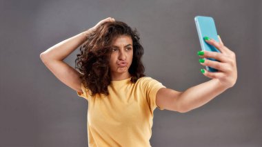 Kıvırcık saçlı hoş bir genç kadın gri arka planda izole edilmiş akıllı telefonuyla selfie çekiyor.