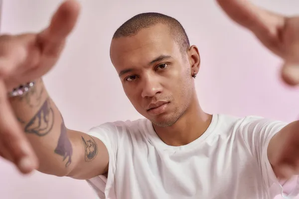 Arca hűvös fiatal vegyes rasszú tetovált srác fehér pólóban nézi a kamera, kinyújtja a karját, miközben pózol elszigetelt rózsaszín háttér — Stock Fotó