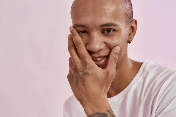 Closeup retrato de feliz jovem mestiço homem tatuado em t-shirt branca sorrindo para a câmera, tocando seu rosto, posando isolado sobre fundo rosa — Fotografia de Stock