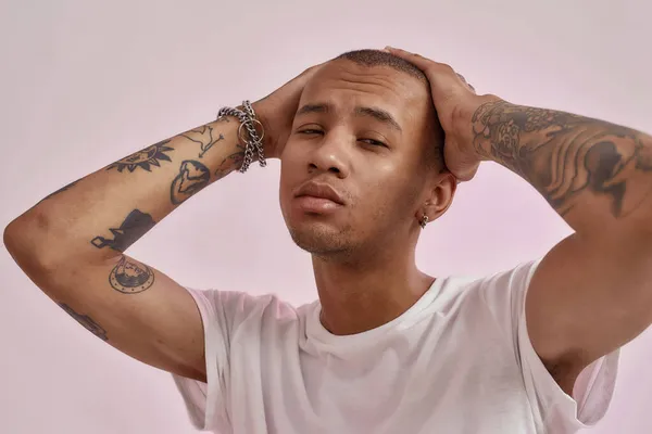 Portré magabiztos fiatal vegyes fajú tetovált fickó fehér pólóban fogja a kezét a fején, miközben pózol a kamera elszigetelt rózsaszín háttér — Stock Fotó
