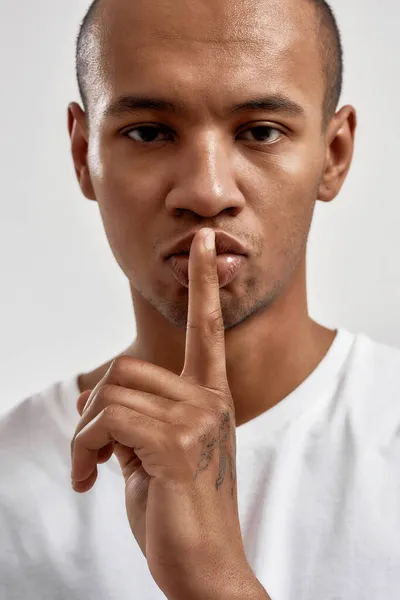 Portrait de jeune homme à la peau foncée montrant un geste shh, gardant un secret, posant isolé sur fond blanc — Photo