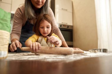 Kız büyükannenin yanında merdaneyle hamuru yuvarlıyor
