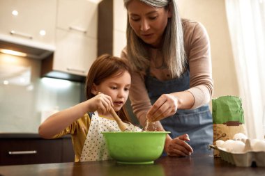 Büyükanne klozete un dökerken, kız hamuru karıştırıyor.
