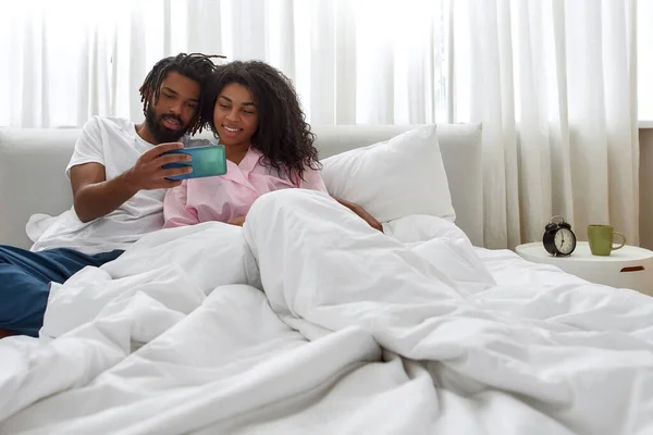 Svart par klocka på smartphone tillsammans på sängen — Stockfoto