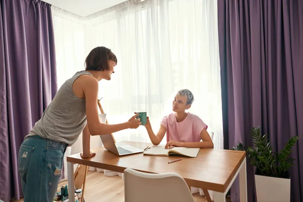 Menina dando chá à namorada que estuda à mesa — Fotografia de Stock